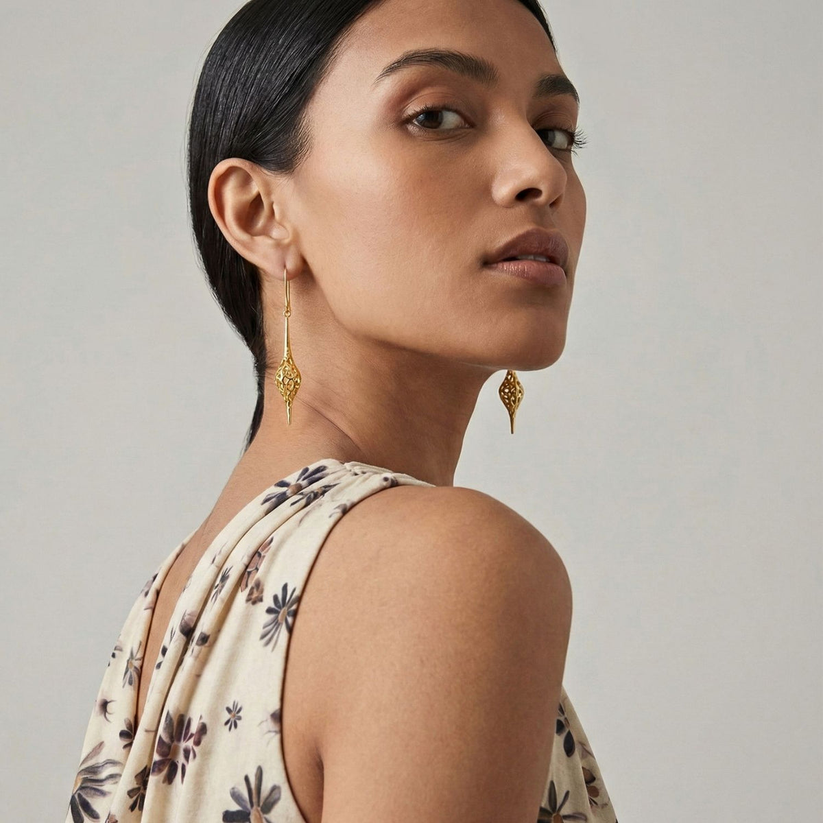 Un woman con dark hair tied back glances over her shoulder, showcasing JAREDJAMIN Joyería Online's oro Jeannie colgante Hook pendientes. Her sleeveless beige, dark-floral top y the neutral background highlight her poised expression y refined joyería.