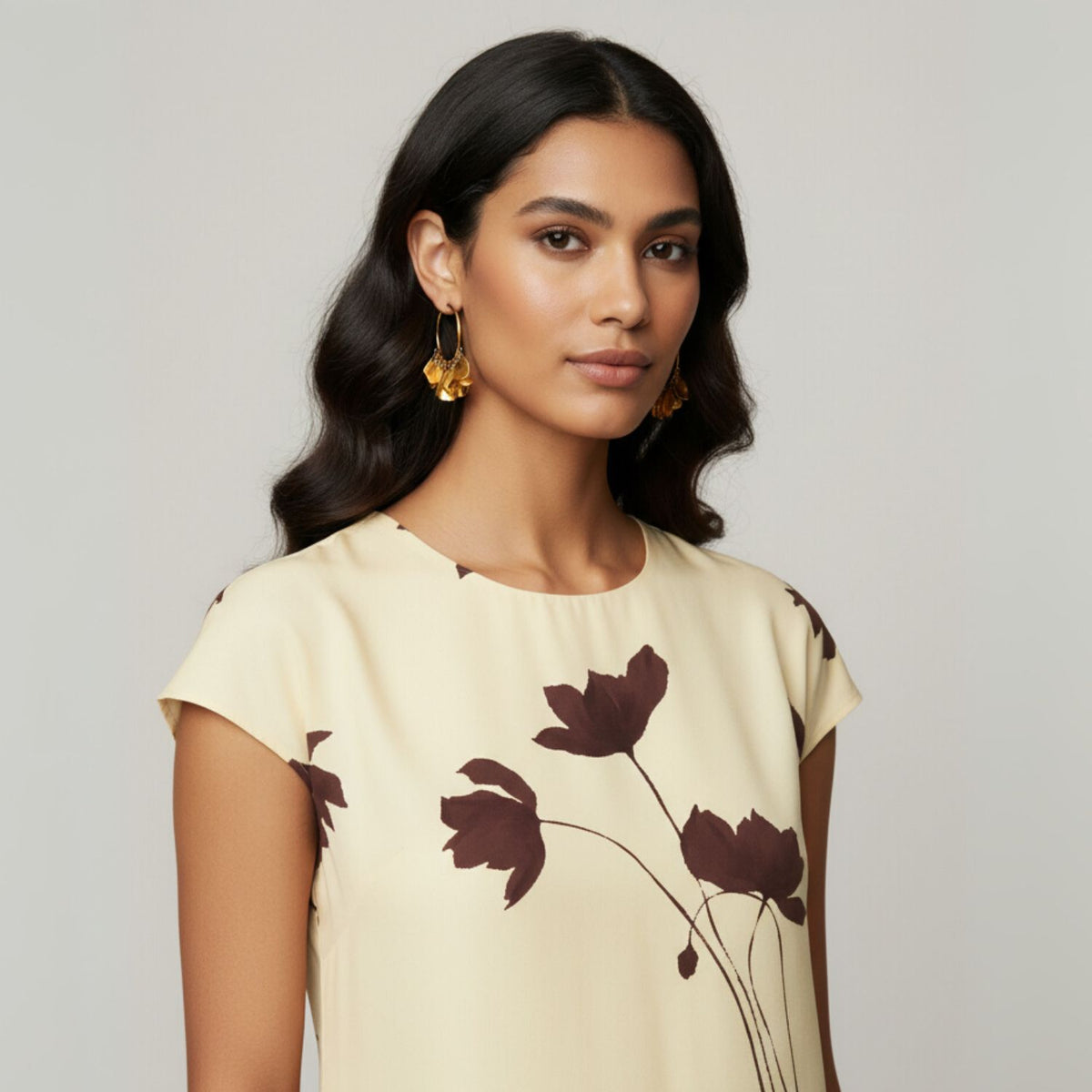 Un woman avec long, wavy dark hair wears JAREDJAMIN Bijoux en Ligne's or Large Petali Click-Top créole boucles d'oreilles et a cream dress avec dark brown floral prints, standing against a plain light gray background et smiling confidently at the camera.