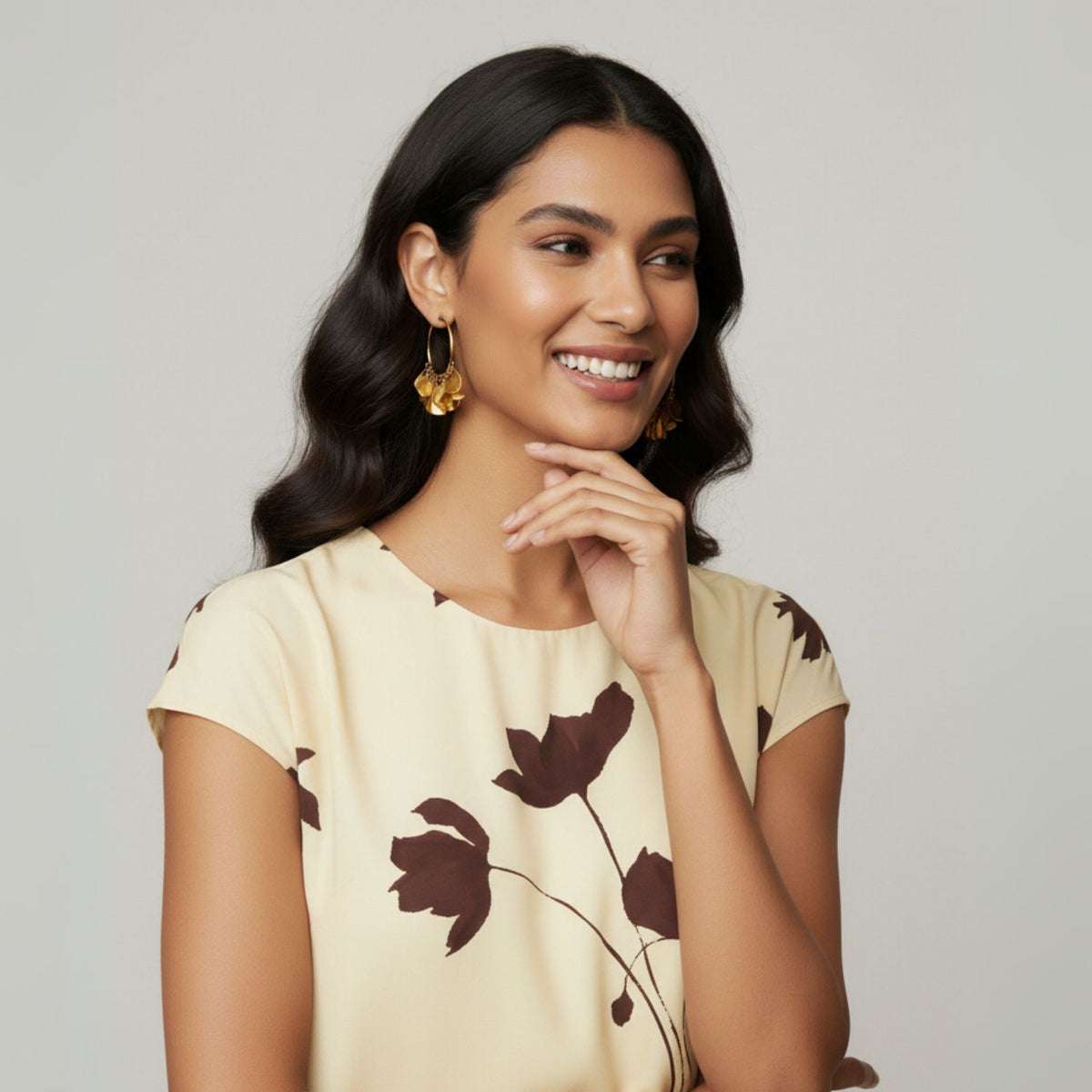 A woman with long, wavy dark hair smiles to the side, wearing JAREDJAMIN Jewelry Online&#39;s Gold Large Petali Click-Top Hoop Earrings and a cream dress with brown floral prints. Her hand is near her chin against a plain light gray background.