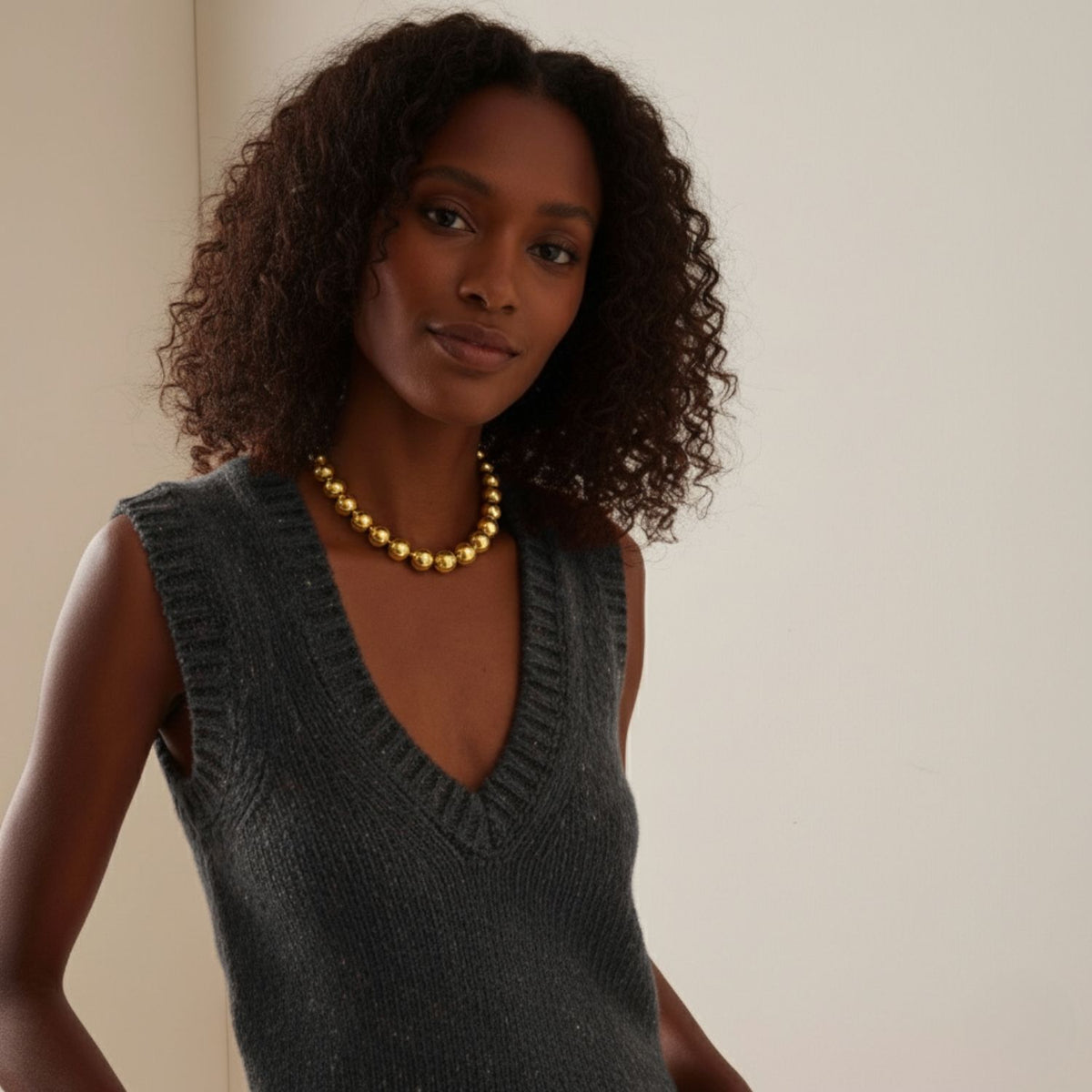 A woman with medium-dark skin and curly hair wears a sleeveless dark gray V-neck sweater and the Lucille Ball Necklace by JAREDJAMIN Jewelry Online. She stands against a light wall, looking at the camera with a slight smile.