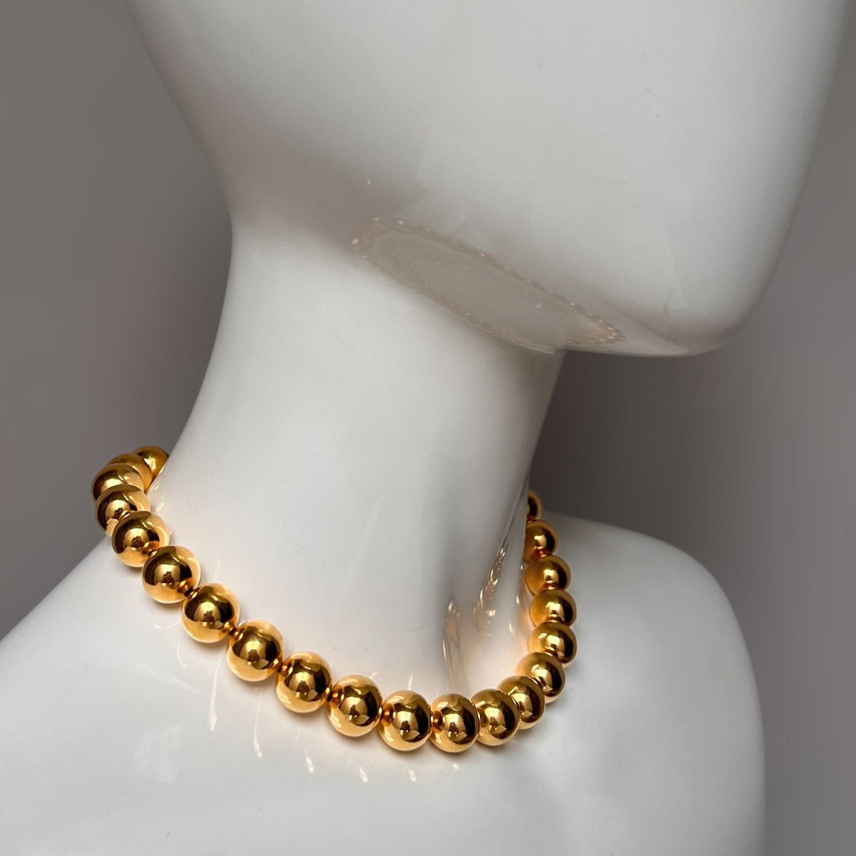 A glossy mannequin showcases the Lucille Ball Necklace by JAREDJAMIN Jewelry Online—large, shiny 18k gold-plated brass beads strung closely and fitting snugly around the neck, gleaming against a plain, neutral backdrop.