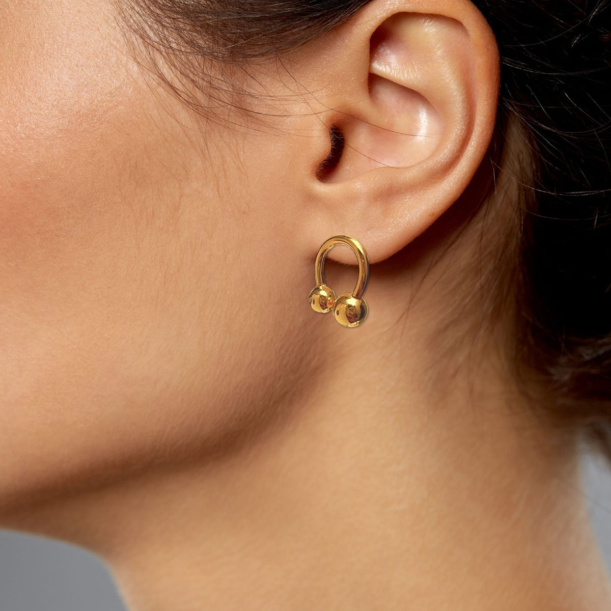 Un gros plan of a woman's ear wearing JAREDJAMIN Bijoux en Ligne's or Lucky Orbita Post boucles d'oreilles, avec brillant 18K or plating et two small or spheres. Her hair is pulled back against a neutral gray background.