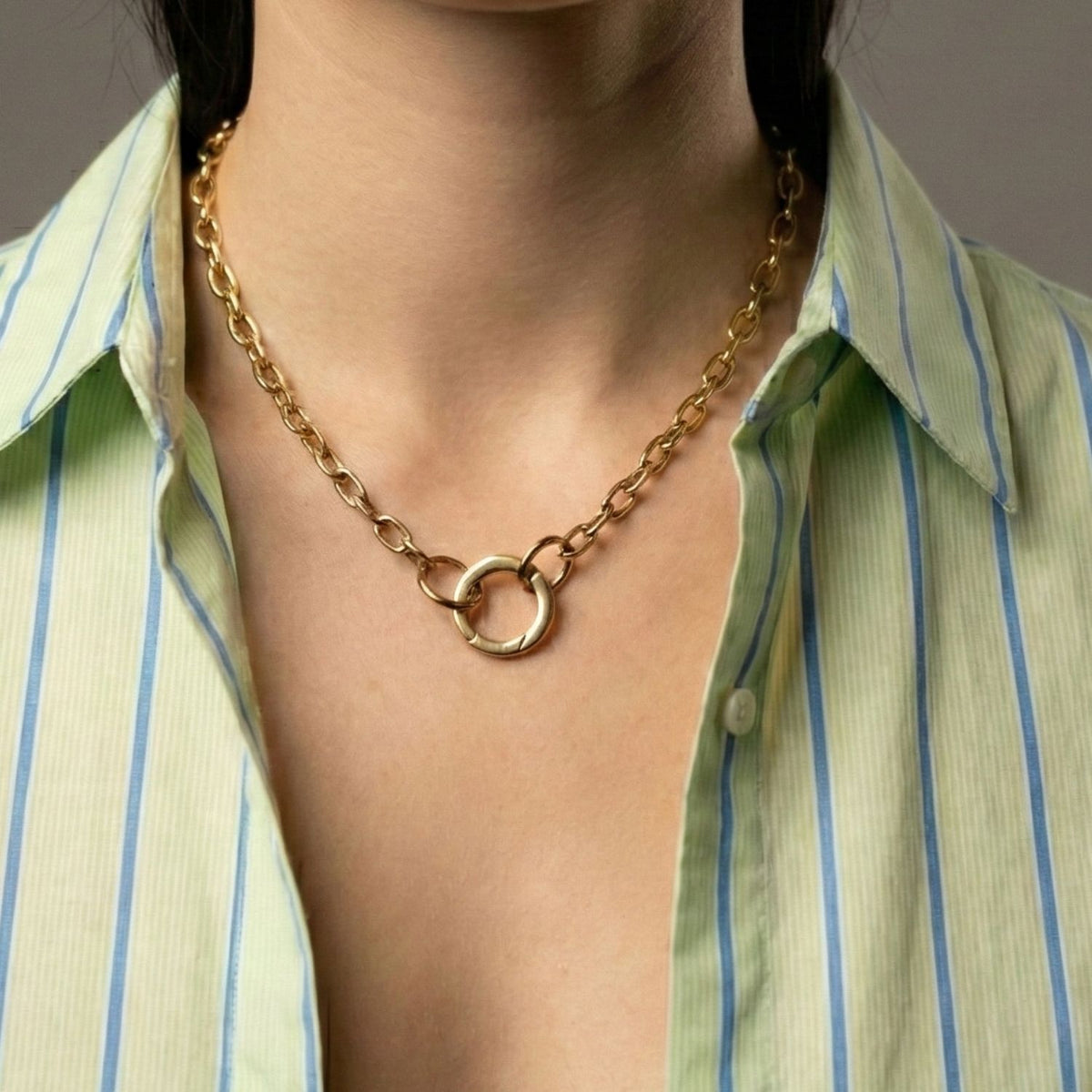 A person in a green and white striped shirt highlights the JAREDJAMIN Jewelry Online Gold Medium Circular Clicker Clasp necklace, featuring two interlocking circular pendants, against a plain gray background emphasizing the jewelry.