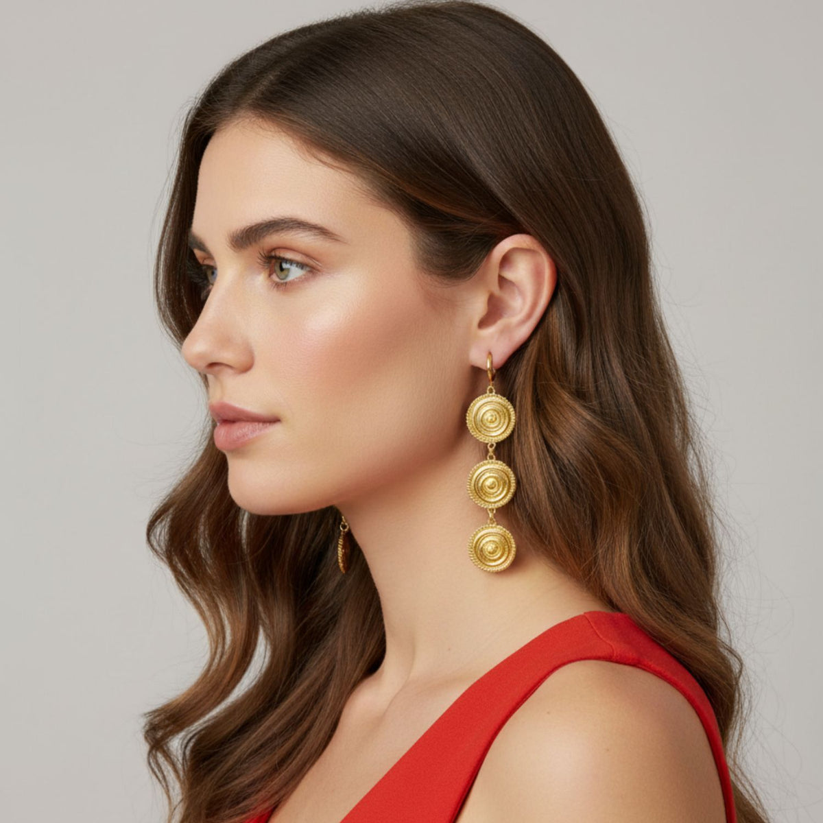 Un woman avec long, wavy brown hair wears a sleeveless red top et JAREDJAMIN Bijoux en Ligne’s or Minerva Leverback boucles d'oreilles—18K plaqué or avec three spiral circles. She faces left against a plain light gray background, her makeup natural et subtle.