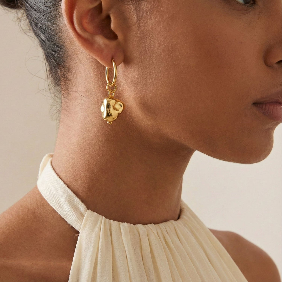 Un woman dans un cream halter-neck top wears JAREDJAMIN Bijoux en Ligne's or Nugget breloque Click-Top créole boucles d'oreilles, avec chunky, abstract breloques. Her hair is tied up et the neutral background highlights her bijoux et outfit.