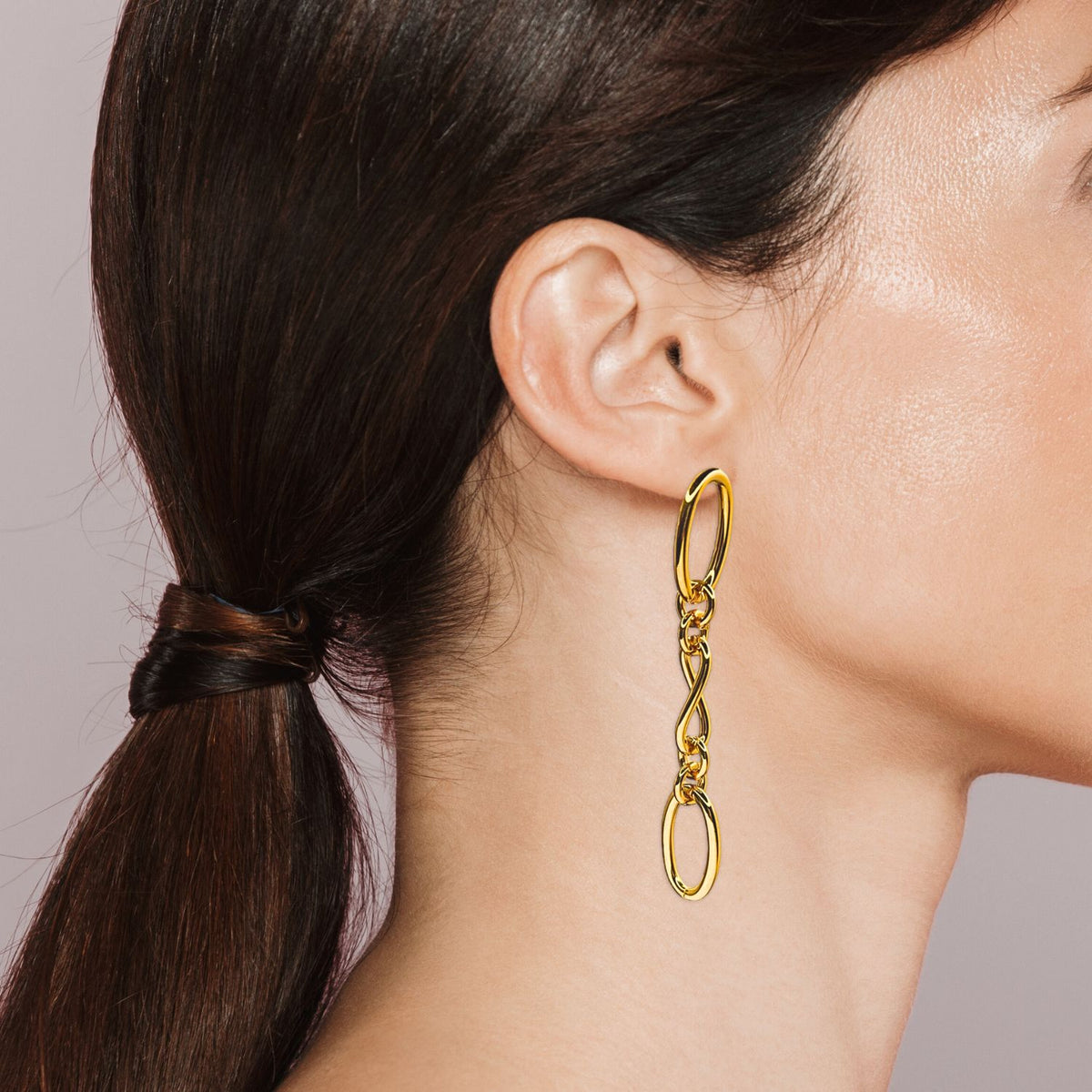 Un woman avec dark brown hair dans un low ponytail wears JAREDJAMIN Bijoux en Ligne’s or Ophelia chaîne Dangle Post boucles d'oreilles. Les plain, light background highlights the boucles d'oreilles et her smooth, slightly tanned complexion.