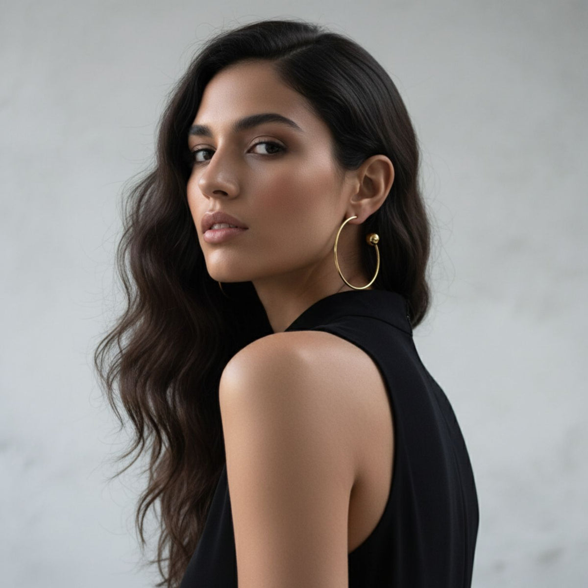 A woman with long, wavy dark hair and light skin wears a sleeveless black top and striking Gold Orbita Comet Earrings by JAREDJAMIN Jewelry Online, looking over her shoulder against a simple light gray background.