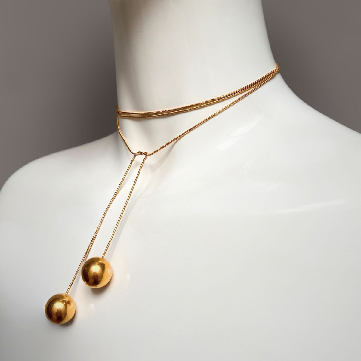 The Gold Orbita Lariat Necklace by JAREDJAMIN Jewelry Online is elegantly displayed on a mannequin, featuring an 18K gold minimalist design with intertwined chains ending in spherical pendants, set against a soft gray gradient background.