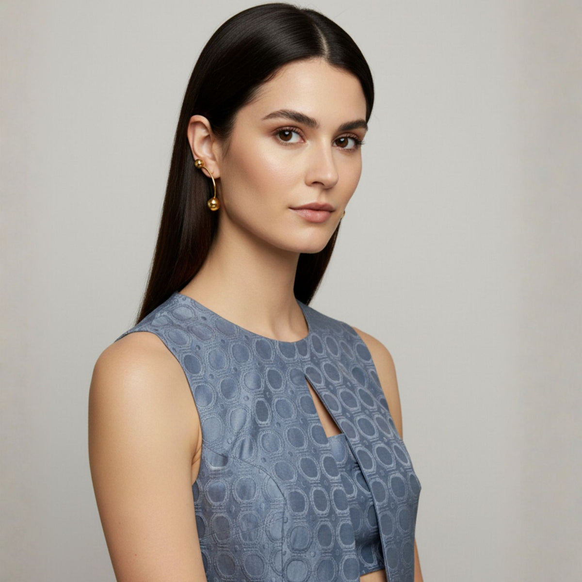 A young womanとlong, straight dark hairとlight skin wears a sleeveless ブルー patterned top 付きの front cutout. She models JAREDJAMIN ジュエリーオンライン ゴールド Orbita Nova Post イヤリング, posing against a plain light background.
