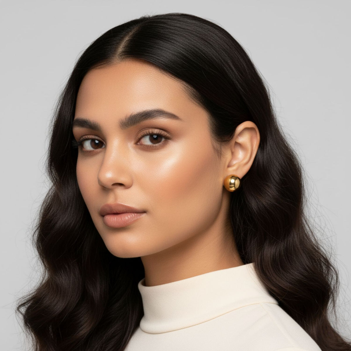 Un woman avec long, wavy dark hair et glowing skin wears a cream turtleneck et JAREDJAMIN or Orbita puce Post boucles d'oreilles, gazing calmly at the camera avec soft makeup against a neutral background, radiating elegance.