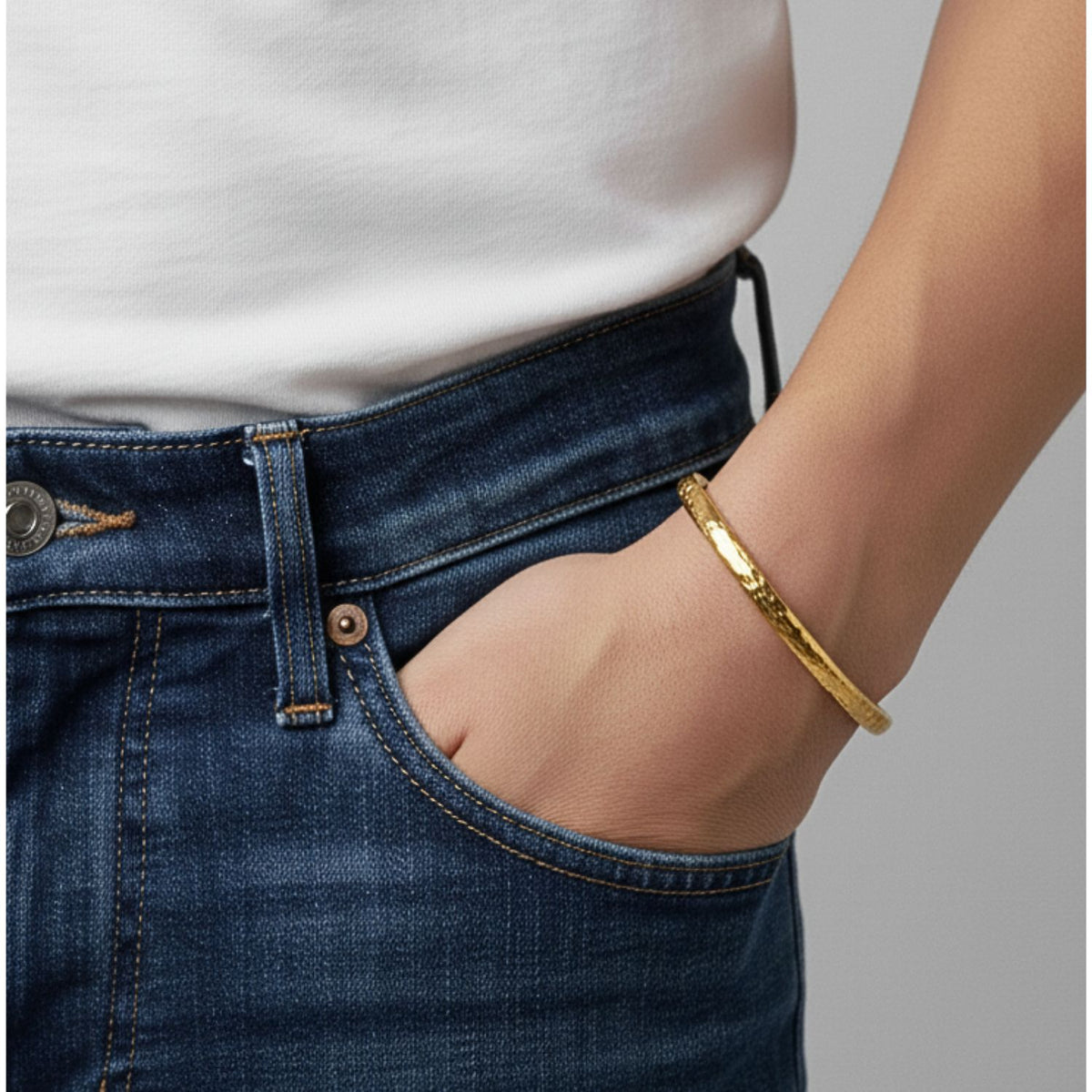 A person in a plain white shirt and blue jeans stands with their right hand in their pocket, showcasing the JAREDJAMIN Jewelry Online Gold Parker Textured Cuff, featuring a hand-hammered finish. The background is neutral and blurred.