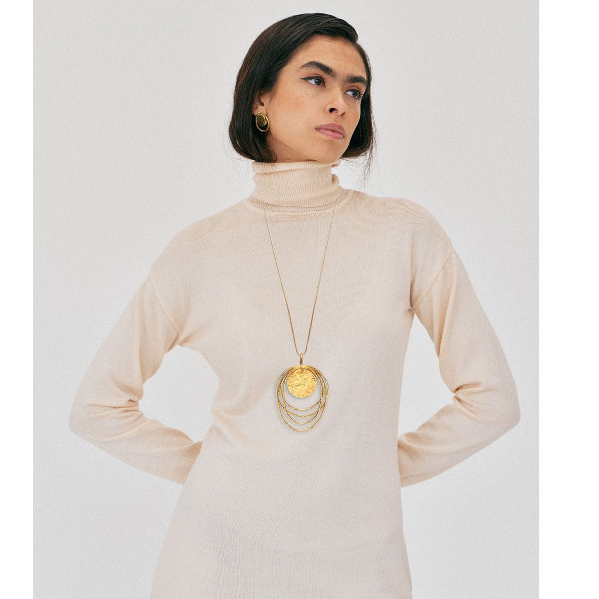 A woman with long dark hair wears a cream turtleneck and gold hoops, looking to the side. She showcases the JAREDJAMIN Jewelry Online Peacock Dawn Gold Pendant Chain Necklace against a plain white background.