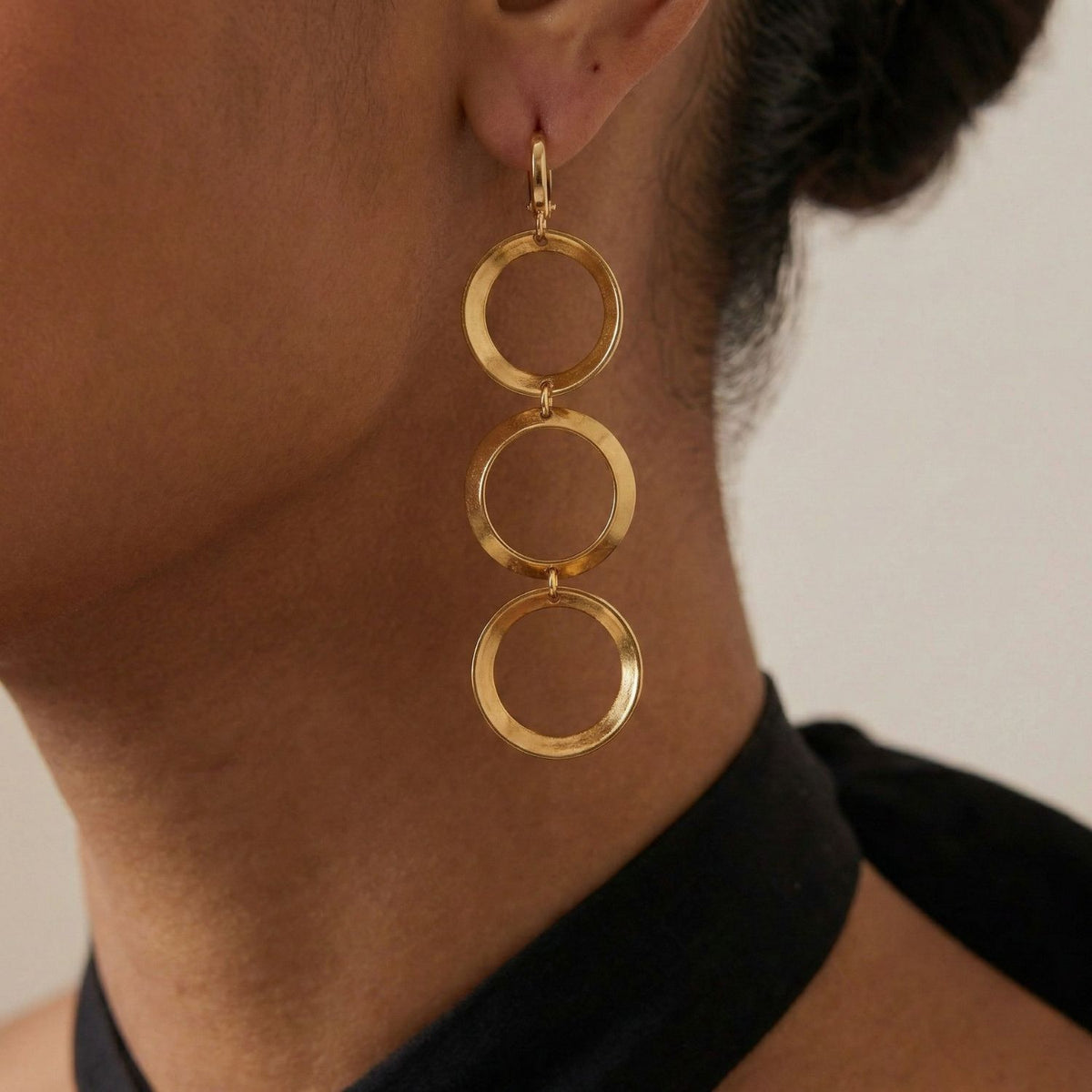 gros plan of a person wearing the JAREDJAMIN Bijoux en Ligne or Rimini Loop boucles d'oreilles—18K or plated avec three linked, brillant open circles. They have medium brown skin, dark hair pulled back, a black halter-top, et a neutral background.