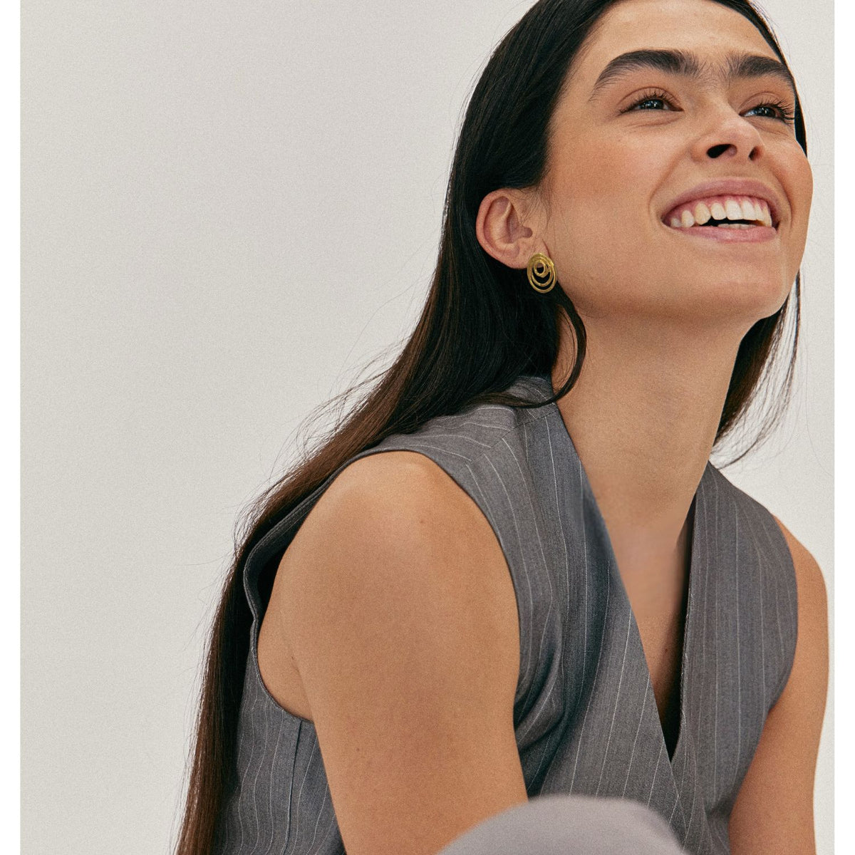 A woman with long dark hair and thick eyebrows is smiling and looking up, wearing JAREDJAMIN Jewelry Online&#39;s Gold Ripple Textured Simple Clip On Earrings. She sits against a plain light background in a closely cropped shot.