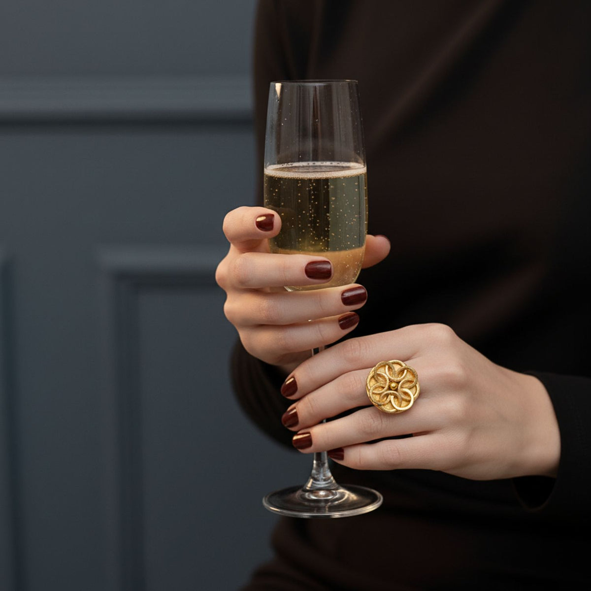 With dark red nails and dressed in black, a person stands against a dark blue paneled wall, holding a champagne flute and wearing the bold, handmade JAREDJAMIN Jewelry Online Gold Santorini Rose Ring on one hand.
