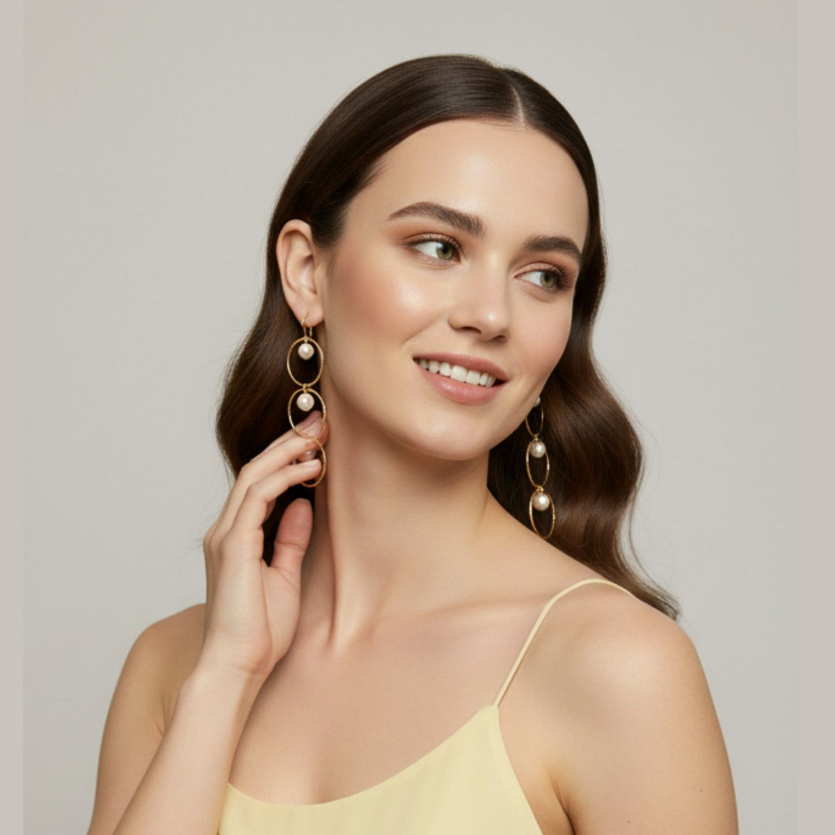 Un woman con long brown wavy hair, smiling y looking left, touches a oro Seabreeze Lindy Dangle pendiente de JAREDJAMIN Joyería Online. She wears a pale yellow spaghetti strap dress against a plain, neutral background.