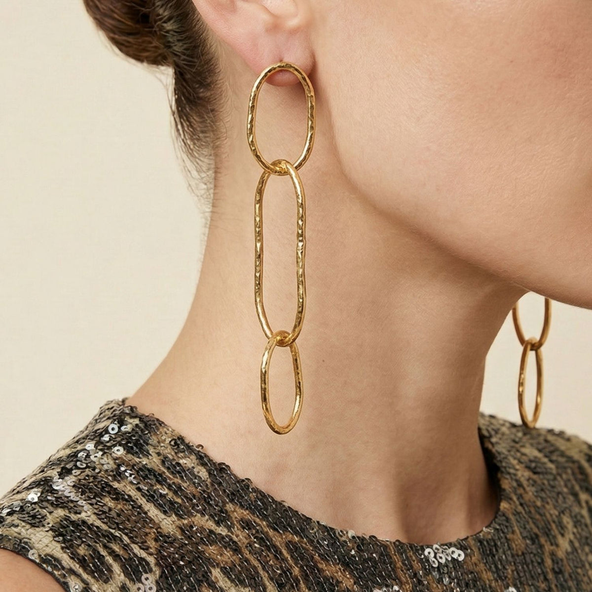 primer plano of a woman's ear wearing JAREDJAMIN Joyería Online's oro Shackle Link colgante pendientes con hammered texture. Her hair is tied back y she wears a sleeveless, sequin animal-print top against a neutral background—luxurious llamativo style.