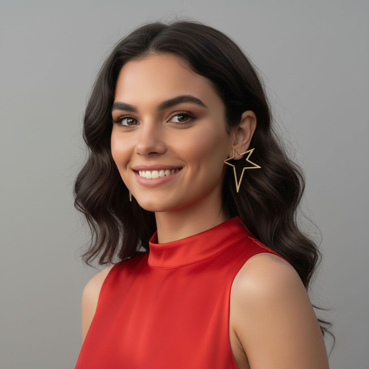 Un woman avec wavy dark hair smiles dans un sleeveless red top, wearing JAREDJAMIN Bijoux en Ligne's or Shooting Star Dust Post boucles d'oreilles. She has natural makeup et poses against a neutral gray background, creating a friendly, élégant look.