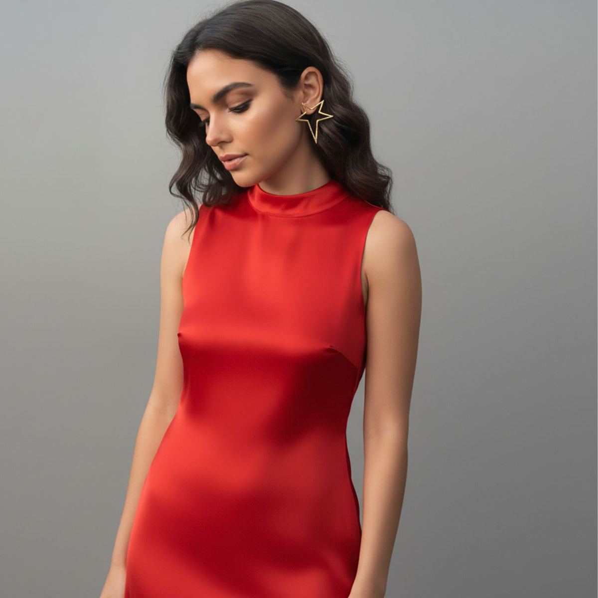 Un woman dans un sleeveless red dress stands against a gray background, wearing wavy dark hair et JAREDJAMIN Bijoux en Ligne's or Shooting Star Dust Post boucles d'oreilles, gazing downward avec a calm, Emily Swallow-like expression.