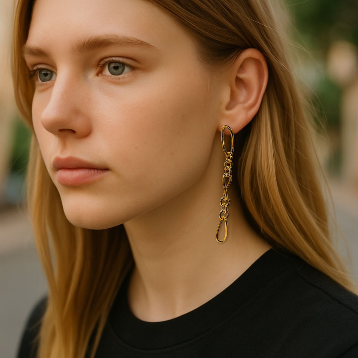 Un young woman avec long blonde hair wears a black shirt et the or Skyler chaîne Post boucles d'oreilles de JAREDJAMIN Bijoux en Ligne. Outdoors, she gazes to the side as her face et boucles d'oreilles stand out against a softly blurred background.