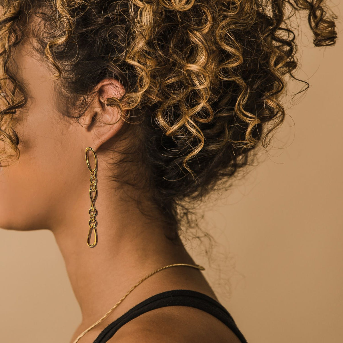 A woman with light brown, curly hair in profile against a beige background wears a thin gold necklace, black top, and JAREDJAMIN Jewelry Online&#39;s Gold Skyler Chain Post Earrings, her face turned away to showcase the stylish jewelry.