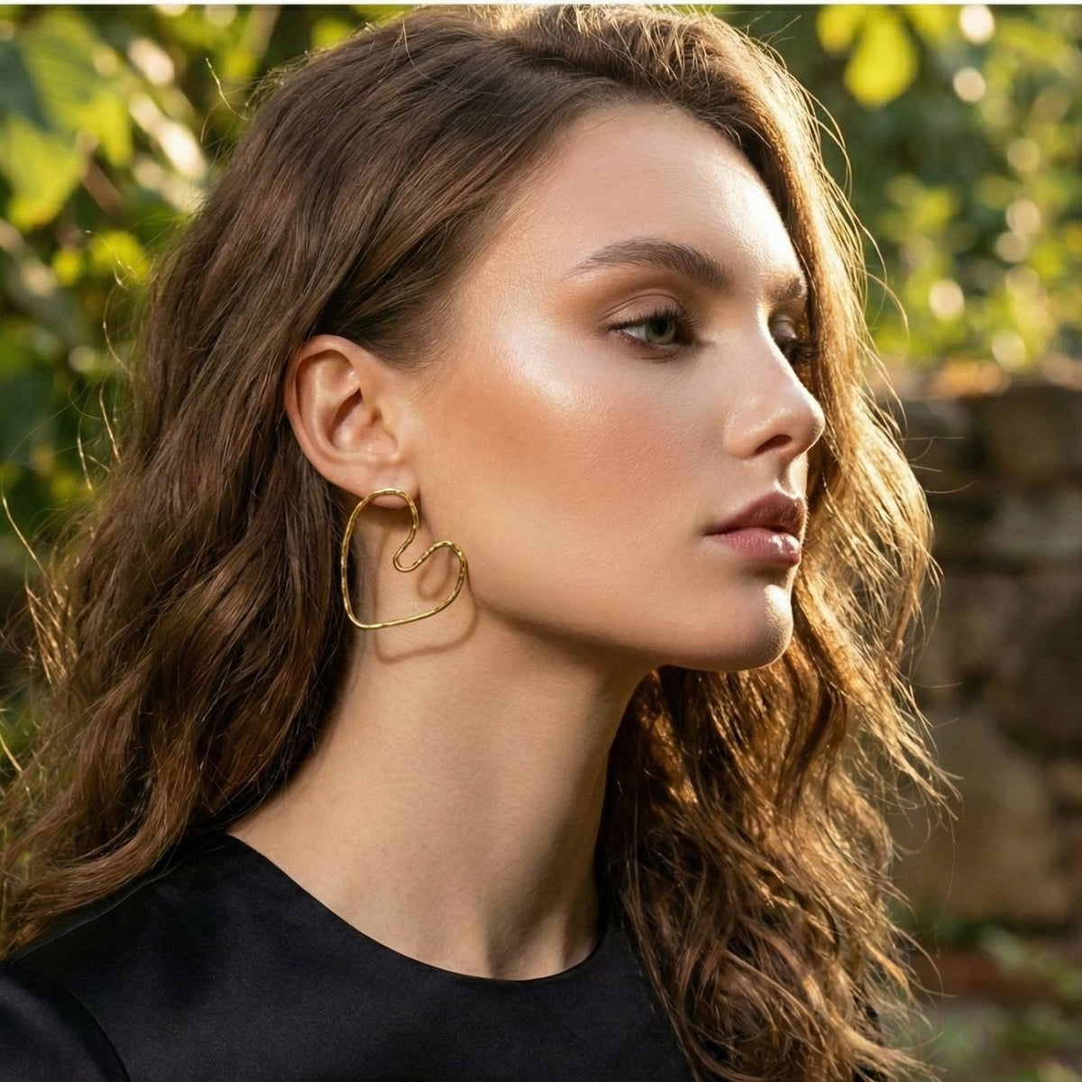 A woman with wavy brown hair, in a black top, models JAREDJAMIN Jewelry Online's Gold Small Fluttering Heart Earrings. She has glowing natural makeup and looks right, outdoors with blurred greenery and sunlight behind her.