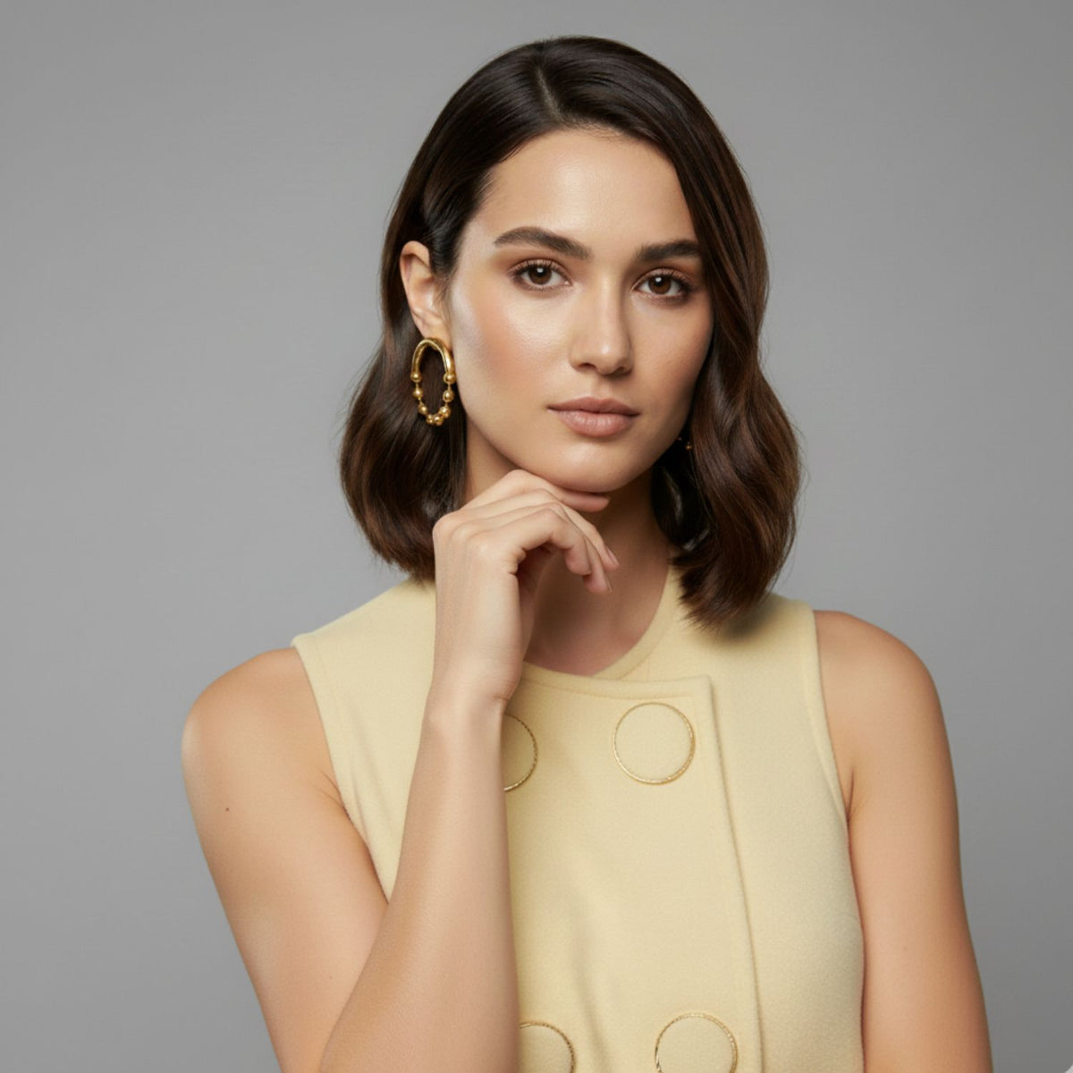 Un woman avec chin-length brown hair dans un sleeveless beige dress avec large buttons wears JAREDJAMIN Bijoux en Ligne's or Small Orbita Amari Post boucles d'oreilles, posing thoughtfully against a gray background avec her hand near her chin et a neutral expression.
