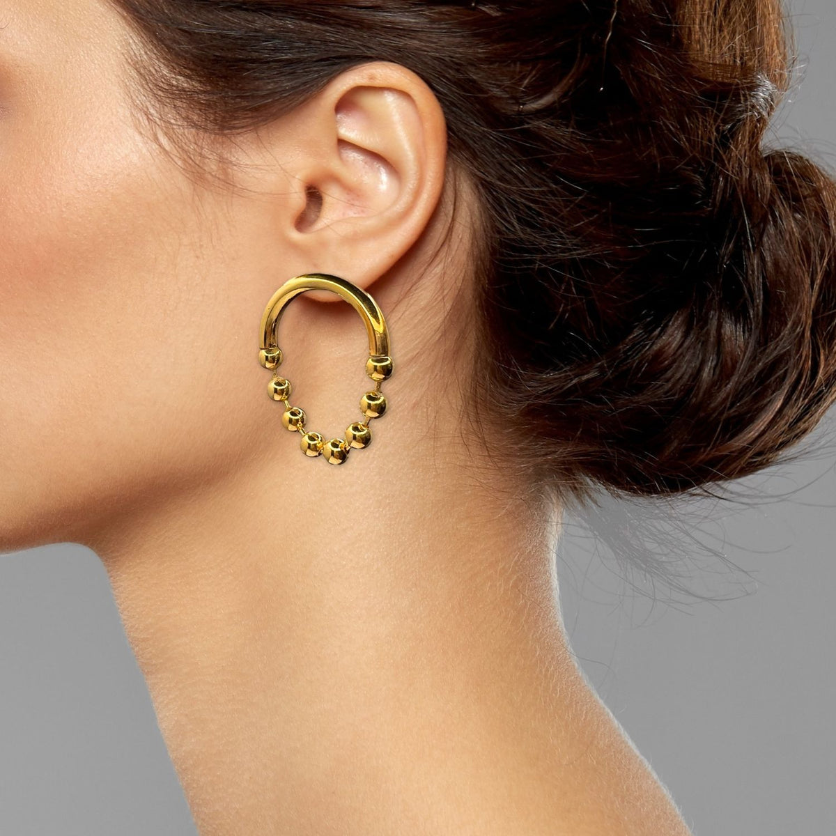A woman with brown hair in a low updo models the Gold Small Orbita Amari Post Earrings by JAREDJAMIN Jewelry Online. The elegant, beaded 18K gold-plated piece stands out against the plain gray background in her side profile.