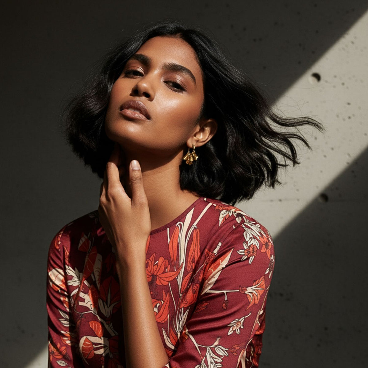 Un woman avec wavy black hair wears JAREDJAMIN Bijoux en Ligne's Small Large Petali Click-Top créole boucles d'oreilles in 18K or plating, posing confidently dans un red floral top as sunlight highlights her face et the texturé wall behind her.