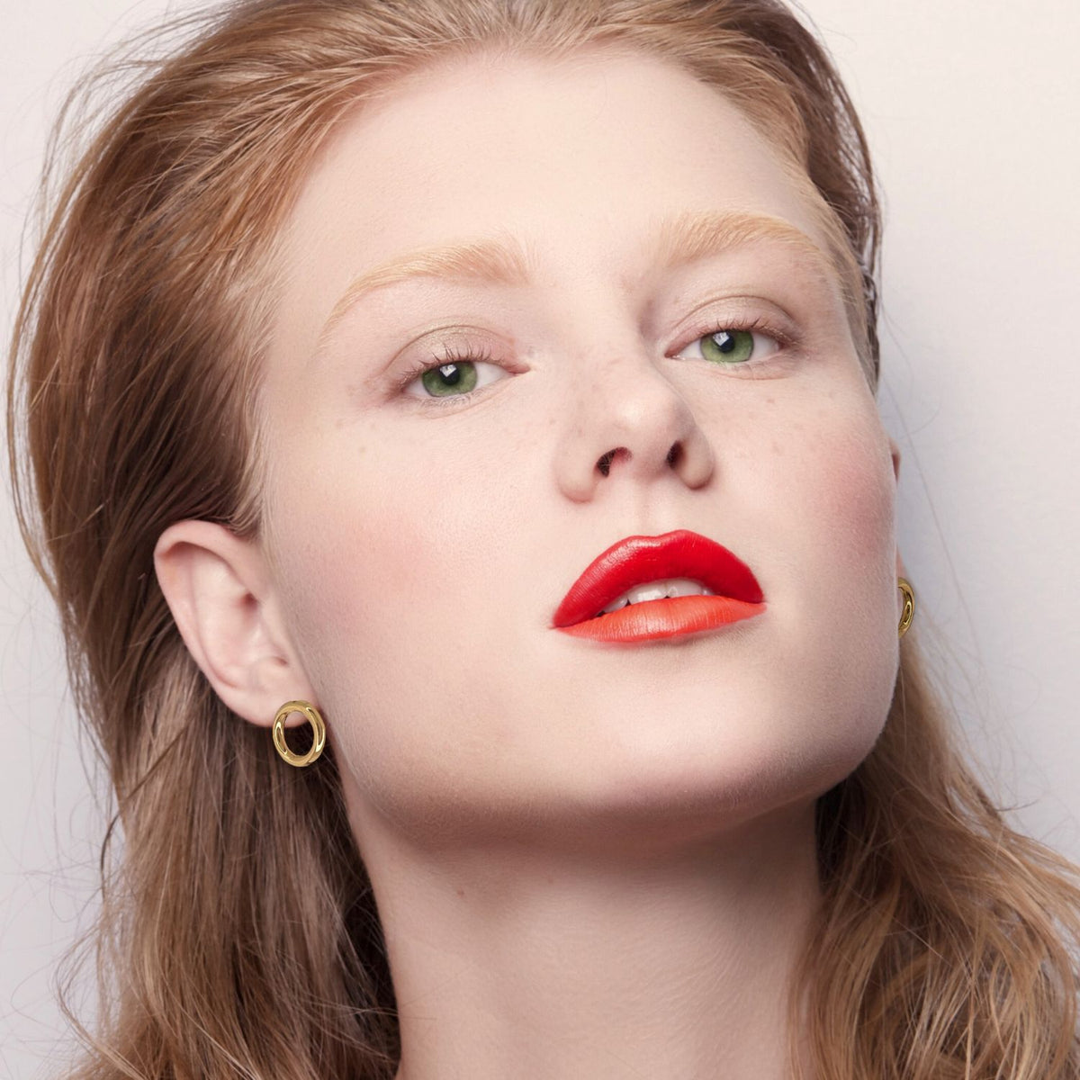 A woman with fair skin, green eyes, and light brown hair looks at the camera, wearing bold red-orange lipstick, subtle blush, minimal eye makeup, and JAREDJAMIN Jewelry Online Gold Small Rhodri Circular Post Earrings against a plain background.