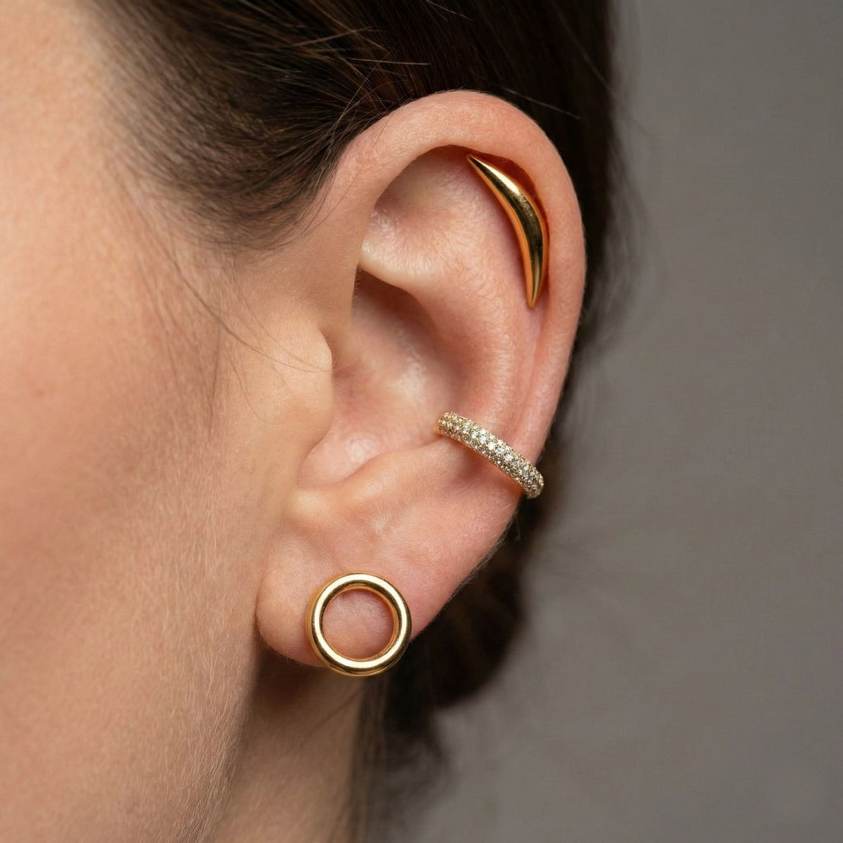 Close-up of a person's ear styled with three earrings: the Elara CZ Gold Plated Ear Cuff by JAREDJAMIN Jewelry Online on the helix, a smooth gold curve on the upper ear, and a simple gold circle loop in the lobe. Plain neutral background.