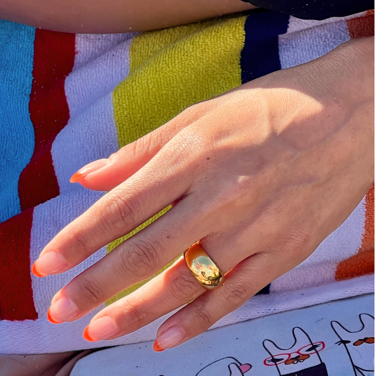 A hand with orange-tipped nails wears the Smooth Dome Ring by JAREDJAMIN Jewelry Online, resting on a colorful striped towel. Sunlight and a peek of a cartoon cat illustration add casual elegance to the scene.