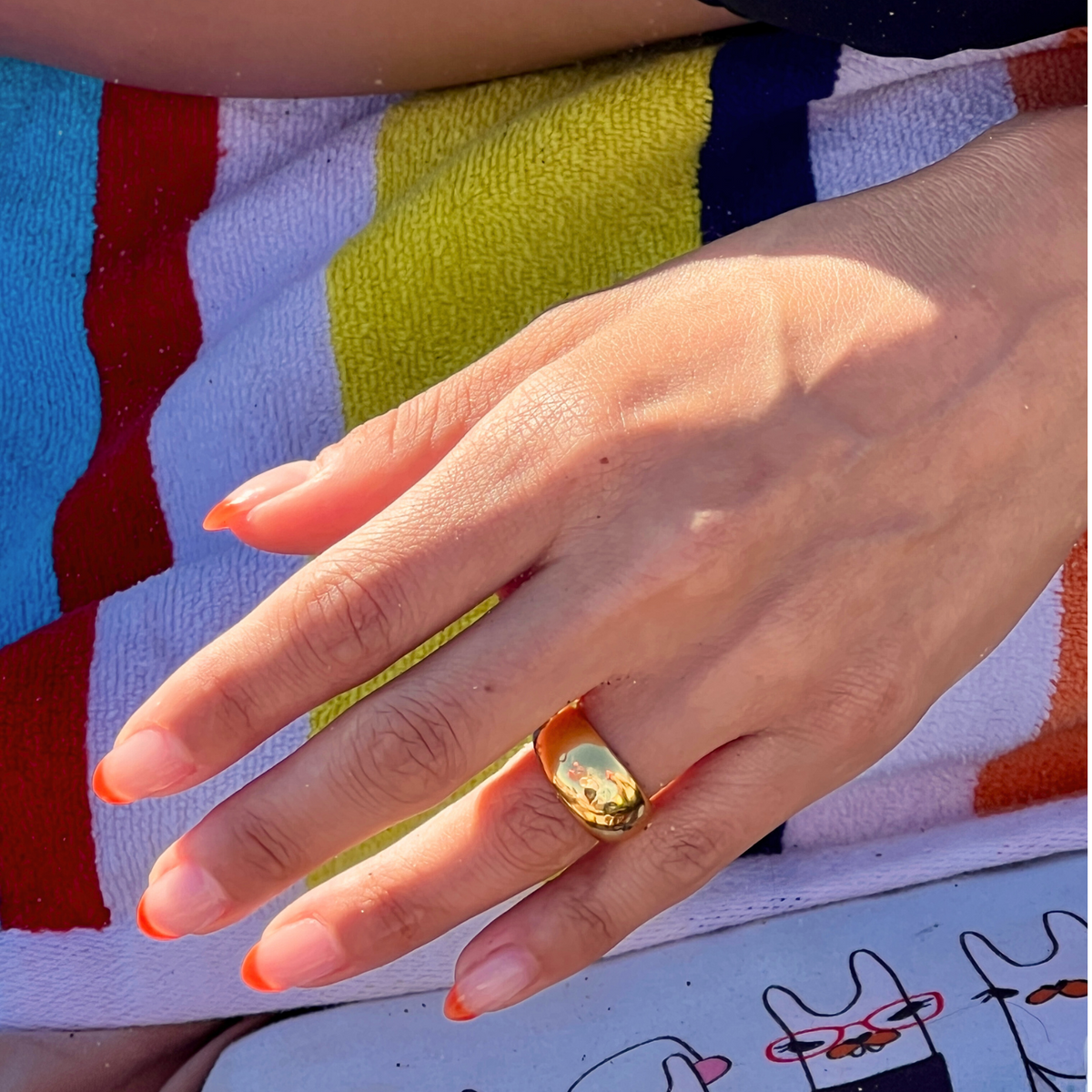 An orange-tipped hand rests on a multicolored towel beside a bag with cartoon cat designs. The Smooth Dome Ring by JAREDJAMIN Jewelry Online gleams in the sunlight, casting an elegant shadow against the vibrant scene.