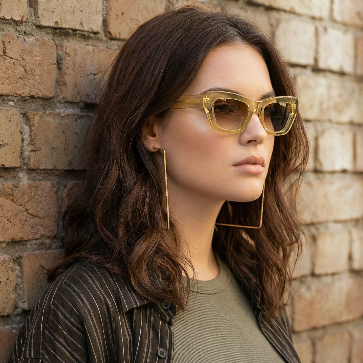 A woman with wavy brown hair stands against a brick wall, wearing square yellow sunglasses and JAREDJAMIN Jewelry Online's Gold Squaretastic Hoop Post Earrings. She styles an olive green top with a dark striped shirt, looking to the side confidently.
