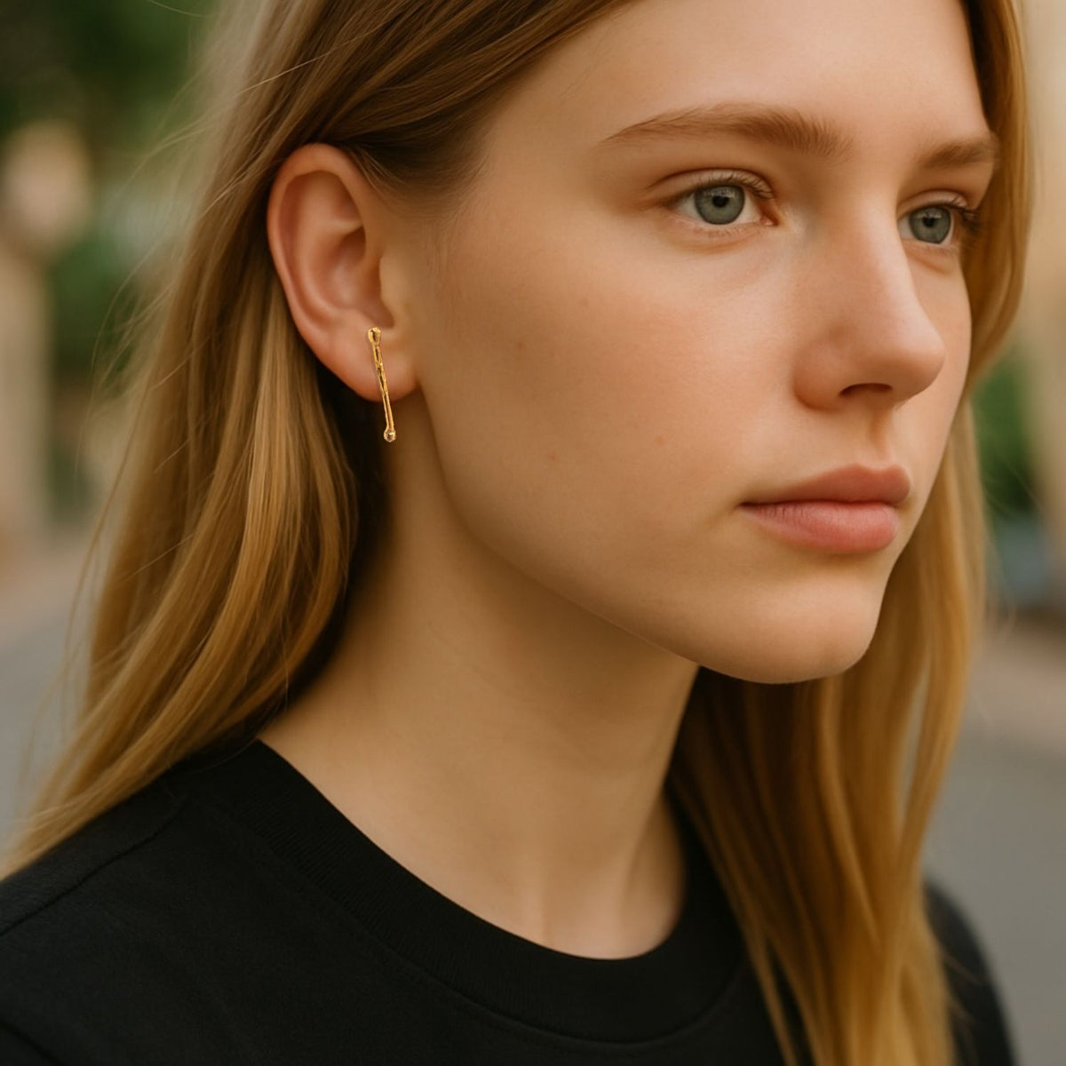 A young woman with long blonde hair and fair skin wears a black shirt and JAREDJAMIN Jewelry Online&#39;s Gold Stix Post Earrings. Outdoors, she gazes to the side with a neutral expression against a softly blurred background.