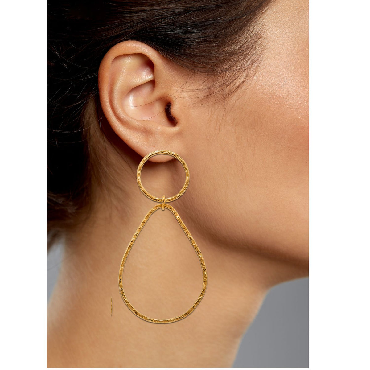 Close-up of a person wearing JAREDJAMIN Jewelry Online&#39;s Gold Textured Tear Post Earrings in 18K gold plating, featuring elongated teardrops on circular hoops with a hammered brass finish against their visible ear and jawline on a plain gray background.