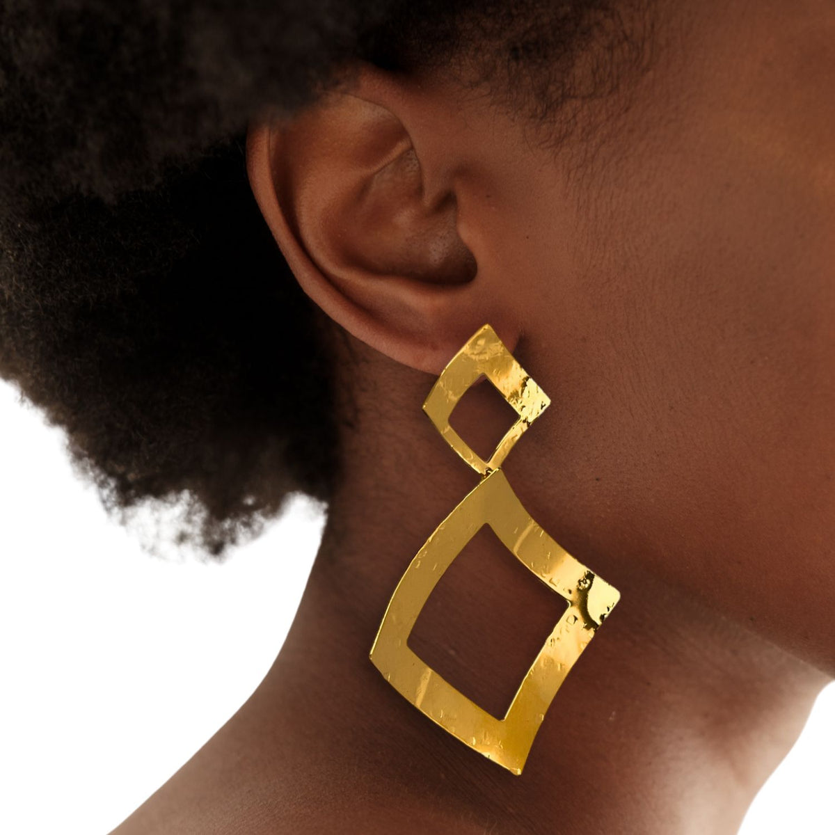 Close-up of a person with dark brown skin and natural curly hair wearing JAREDJAMIN Jewelry Online’s Gold Toscana Diamond Drop Post Earrings—large, abstract 18K gold plated squares—from the Toscana Collection, against a white background.