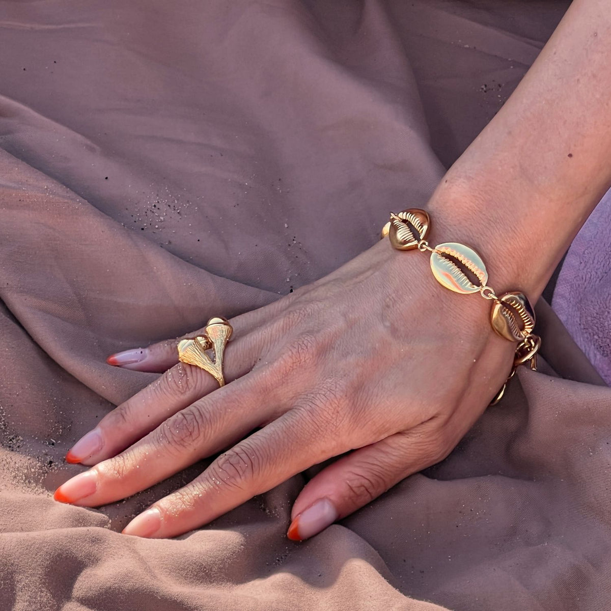A hand with almond-shaped nails fading from nude to orange, dusted with fine sand, rests on mauve fabric. The person wears the JAREDJAMIN Jewelry Online Gold Trinity Acorn Ring and an 18K gold plate bracelet with seashell charms.