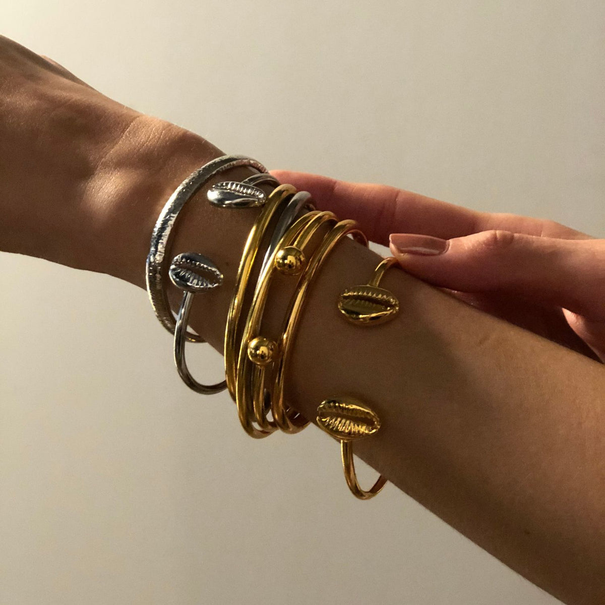 A person’s arm wears the JAREDJAMIN Jewelry Online Silver Tubular Cuff, paired with gold and silver-toned bangles featuring cowrie shell and sphere accents. Another hand gently holds the wrist against a softly lit, plain background.