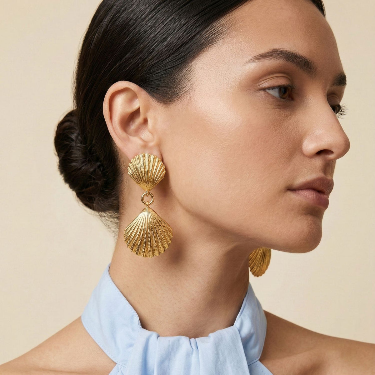 A woman with dark hair in a low bun wears JAREDJAMIN Jewelry Online's Gold Venus Clip On Earrings, featuring 18k gold plating. Her calm expression and light blue halter-neck top enhance the resort-ready style against a neutral backdrop.