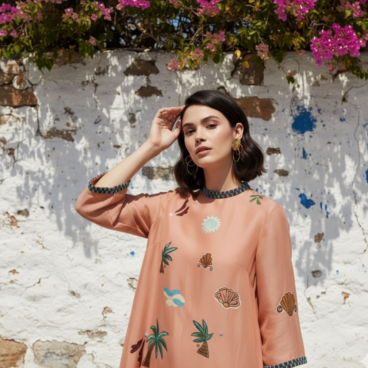 A woman with dark wavy hair in a peach dress with shells, palm trees, and fish stands by a white stone wall and pink flowers, shielding her eyes from the sun and finishing her look with JAREDJAMIN Jewelry Online&#39;s Gold Viviane Clip-on Earrings.