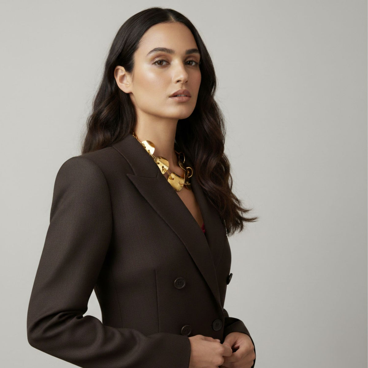A woman with long, wavy dark hair wears a dark brown double-breasted blazer and the Gold Viviane Necklace by JAREDJAMIN Jewelry Online—a chunky, handcrafted piece with a hammered texture. She gazes confidently at the camera against a light gray background.