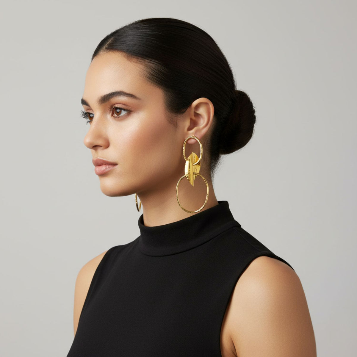 A woman with smooth dark hair in a low bun wears Gold Viviane Post Drop Earrings by JAREDJAMIN Jewelry Online, styled with a sleeveless black top. Shown in profile against a light gray background, she has natural makeup and a composed expression.