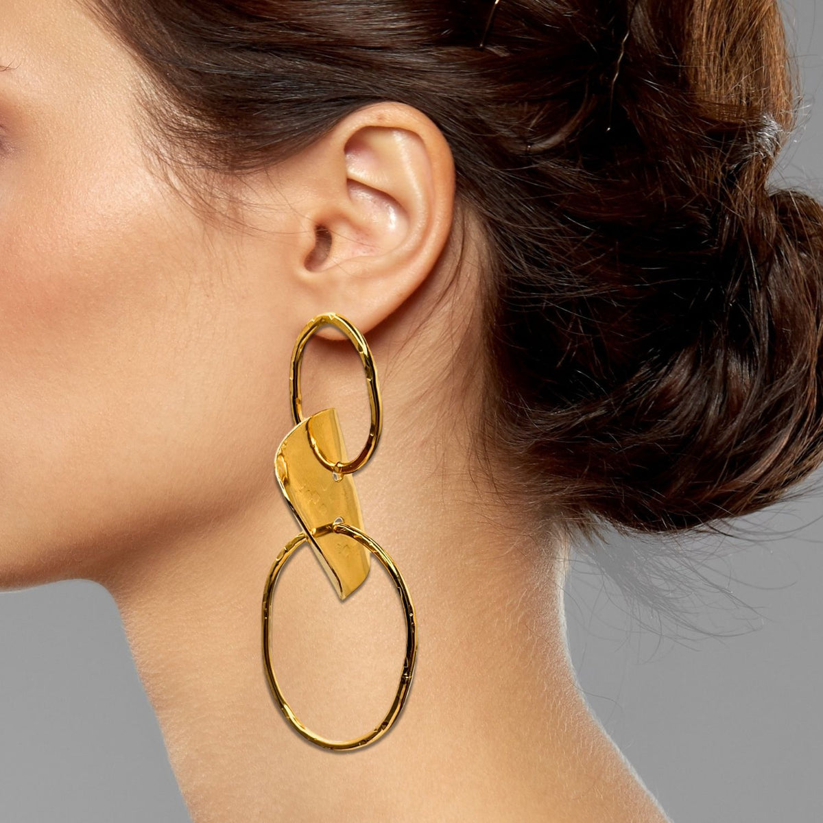 A close-up shows a woman’s dark brown hair in a loose updo as she wears JAREDJAMIN Jewelry Online’s Gold Viviane Post Drop Earrings, featuring bold hoops and hammered metal details. The gray background is plain.
