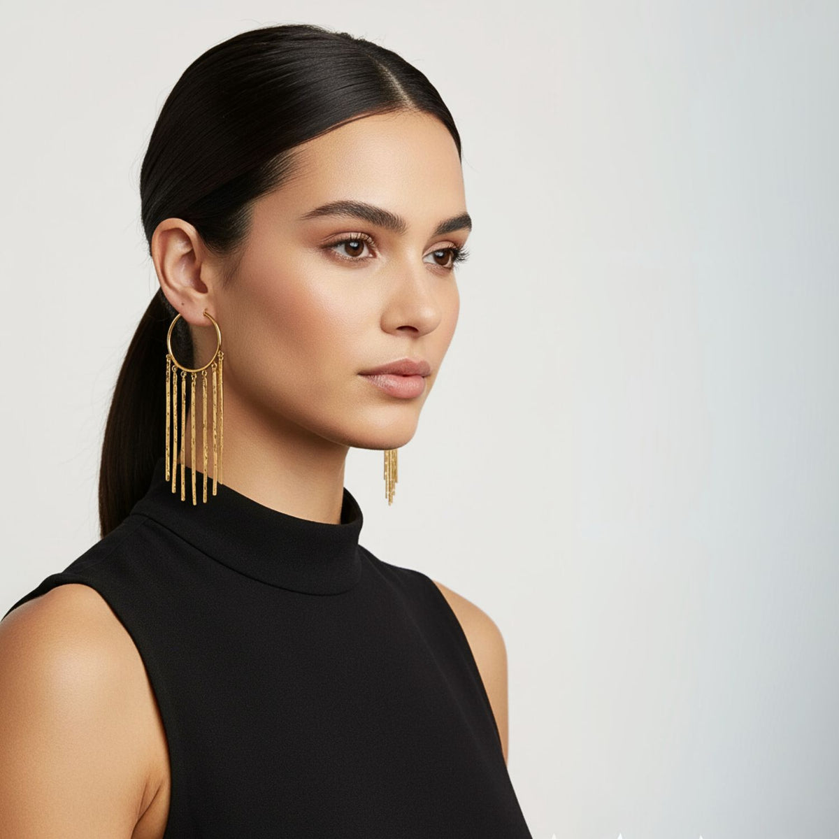 Un woman con sleek dark hair en un low ponytail wears a sleeveless black mock-neck top y JAREDJAMIN Joyería Online’s oro Wind Catcher Leverback Dangle pendientes. She looks to the side con a neutral expression against a plain light background.