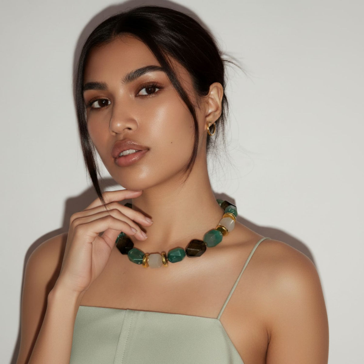 A woman with medium skin and dark hair in a low updo poses against a white background, wearing a pale green spaghetti strap top and the Green Dream Necklace by JAREDJAMIN Jewelry Online, paired with gold hoop earrings and natural makeup.
