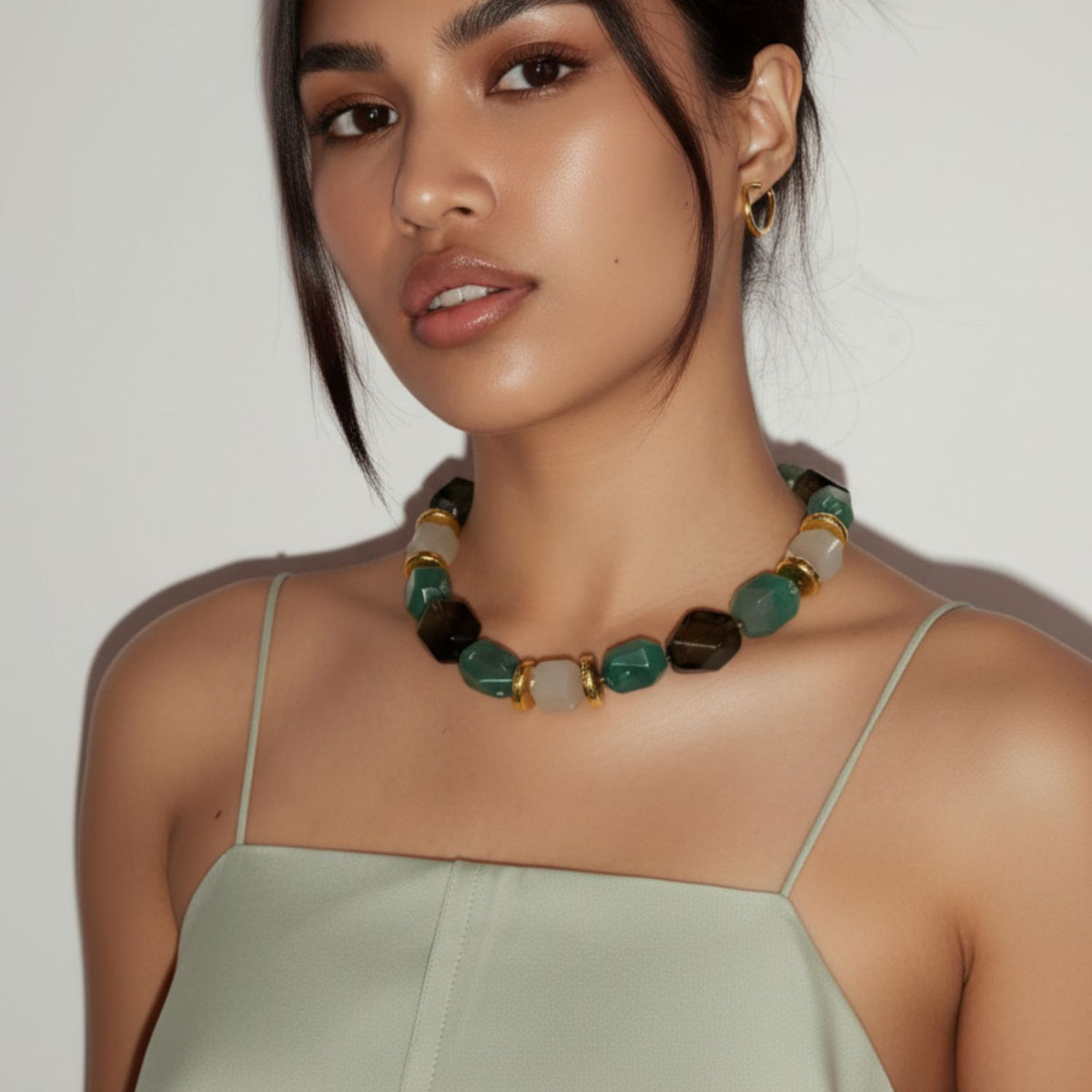 A woman with medium skin tone and dark hair wears gold hoop earrings and the Green Dream Necklace by JAREDJAMIN Jewelry Online—a chunky, hand-knotted statement piece with green, brown, and gold beads. She faces the camera neutrally.