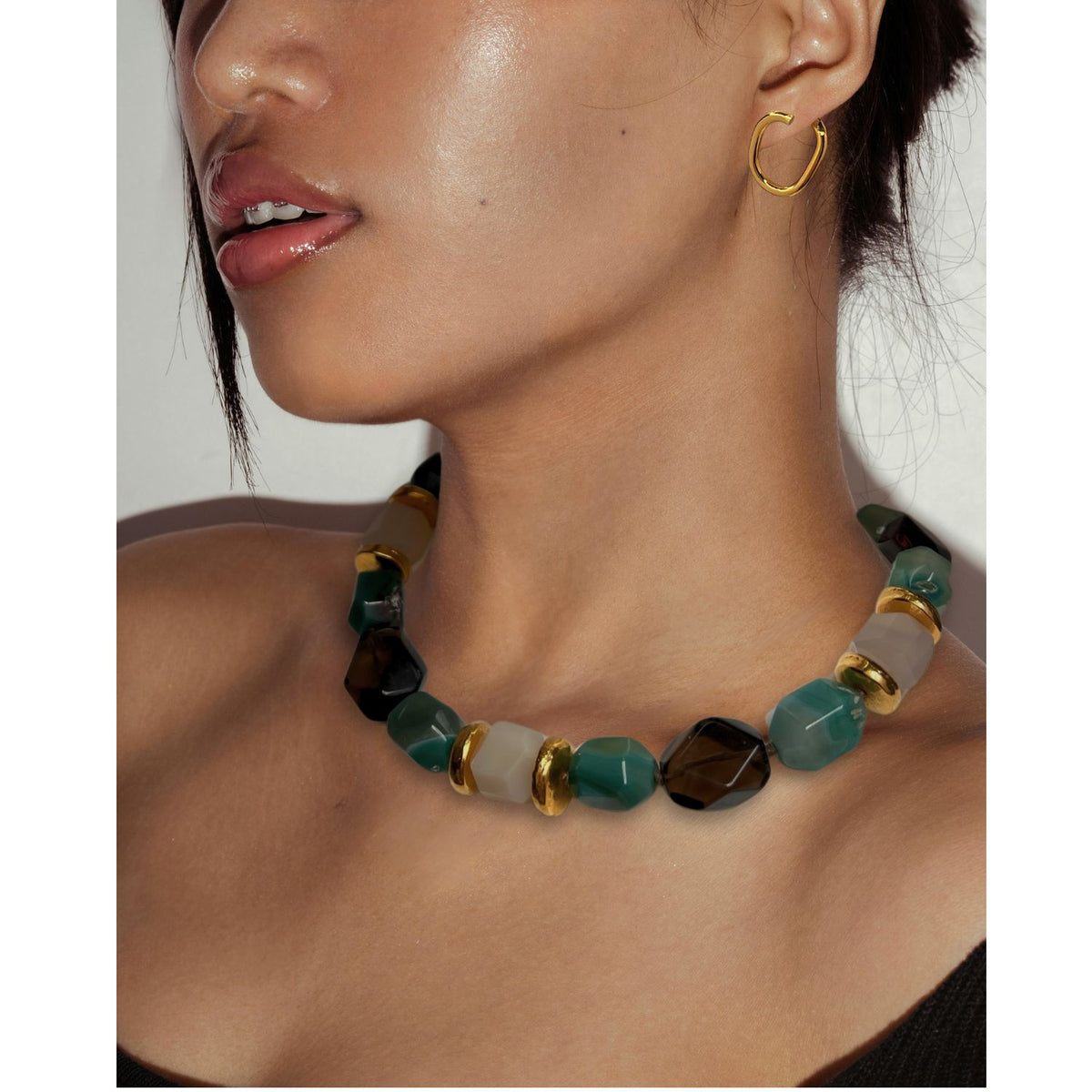 A woman with glossy lips wears the Green Dream Necklace by JAREDJAMIN Jewelry Online—large, geometric beads in green, black, white, and gold with Green Agate. She also wears a small gold hoop earring under soft lighting.