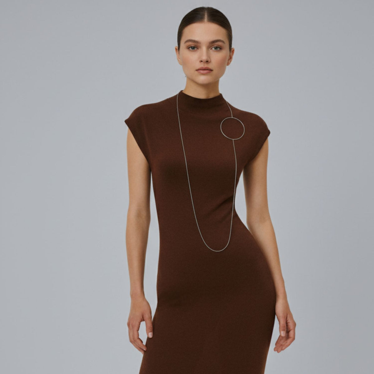 A woman with dark hair tied back wears a fitted brown dress, styling the Silver Halo Necklace by JAREDJAMIN Jewelry Online—a versatile, long toggle clasp piece—while standing confidently against a plain gray background.