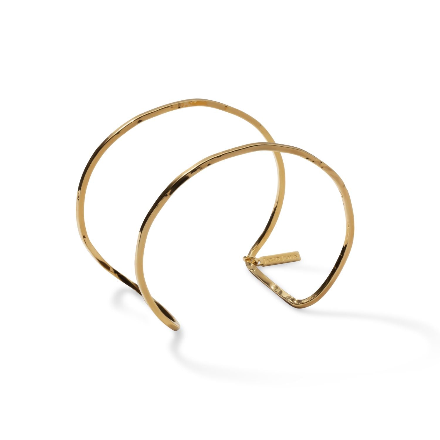 The Gold Halo Forge Textured Cuff by JAREDJAMIN Jewelry Online is a minimalist, open-ended 18K gold-plated brass cuff with a hammered finish, two overlapping curved bands, and a small rectangular tag at one end.